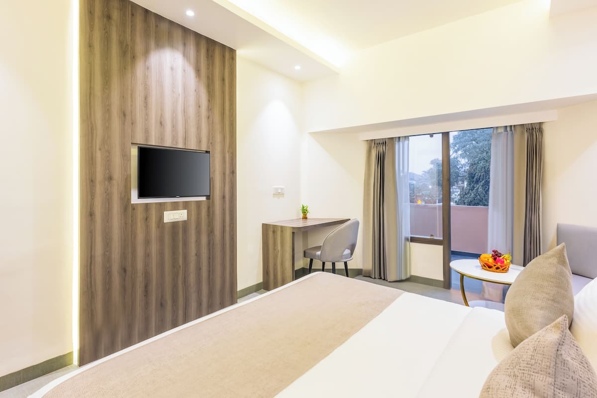 A modern hotel room with a large bed, a desk with a chair, a flat-screen TV mounted on a wooden wall, and a balcony with a view of the city.