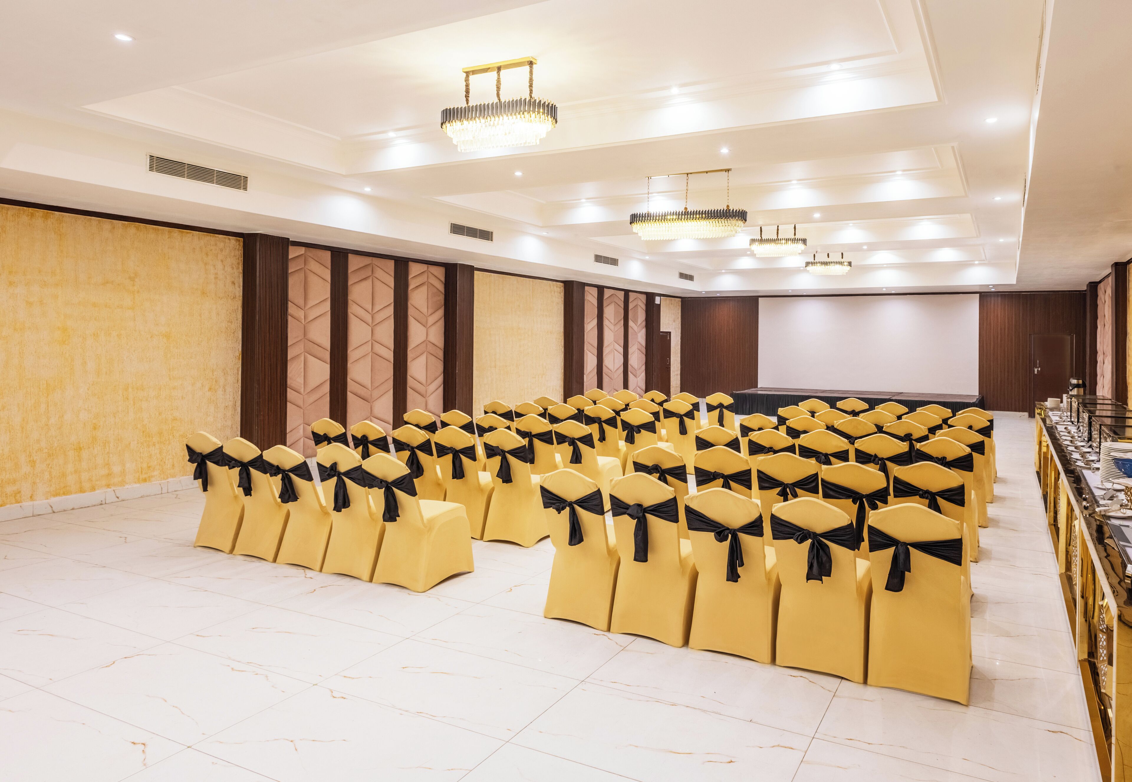 A conference room with rows of yellow chairs and black sashes, a projector screen, and a chandelier.