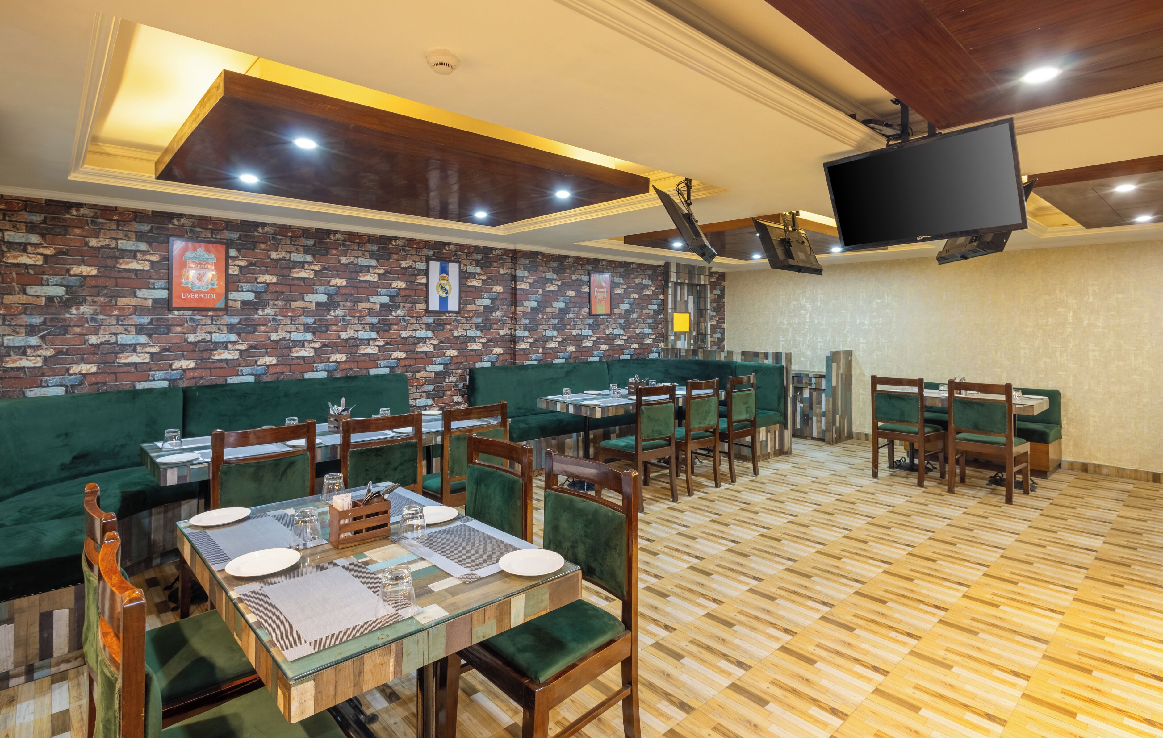 A restaurant with brick-patterned walls, green upholstered booths, wooden tables, and a flat-screen TV mounted on the ceiling.