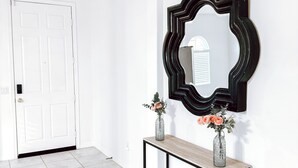 En hall med en vit dörr, ett avlastningsbord med en vas med blommor och en stor spegel på väggen.