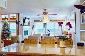 A spacious living room with a large dining table, wicker chairs, a ceiling fan, and a view of the ocean through large windows. - Oceanfront 4BR Home w/ Private Pool, Decks, Beach Walkway, Stunning Views, New to Rental Market (Kure Beach)