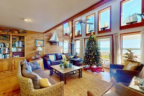 A cozy living room with a large window, a Christmas tree, and a bookshelf filled with books. - Oceanfront 4BR Home w/ Private Pool, Decks, Beach Walkway, Stunning Views, New to Rental Market (Kure Beach)