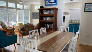 Ein Essbereich mit Holztisch und weißen Stühlen, ein Bücherregal mit Büchern und ein Küchenbereich mit blauen Schränken.