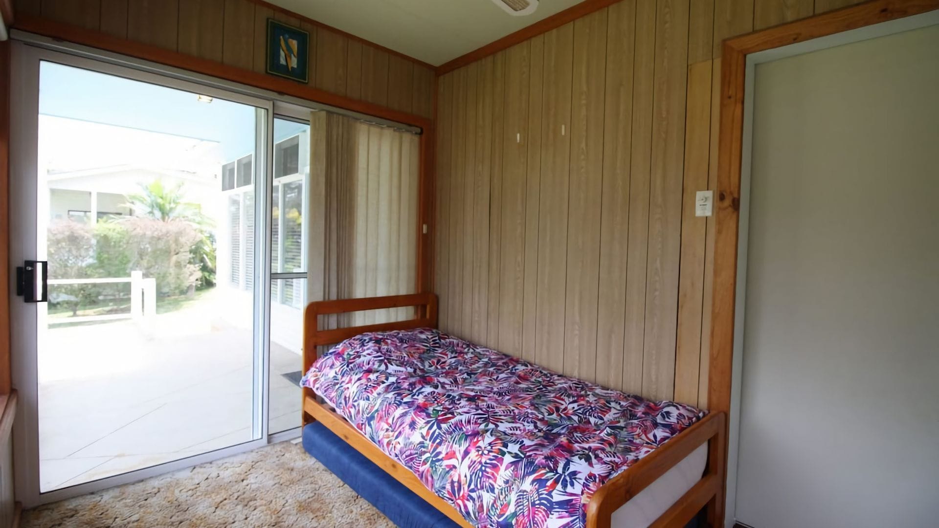 A bedroom with a single bed, a sliding glass door leading to a balcony, and wooden paneling on the walls.