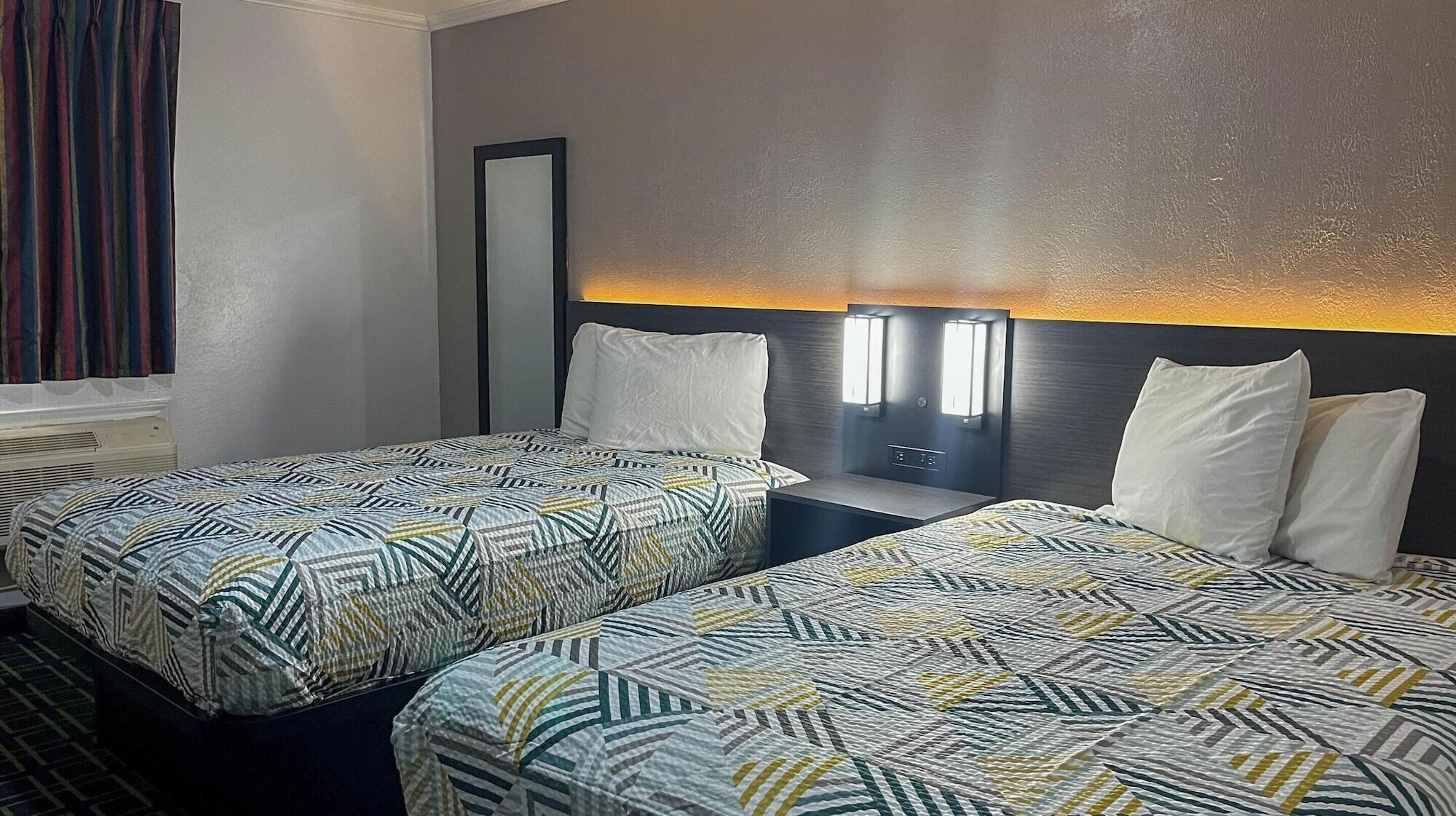 A hotel room with two beds, each with patterned bedding, a nightstand, a mirror, and a wall-mounted light fixture.