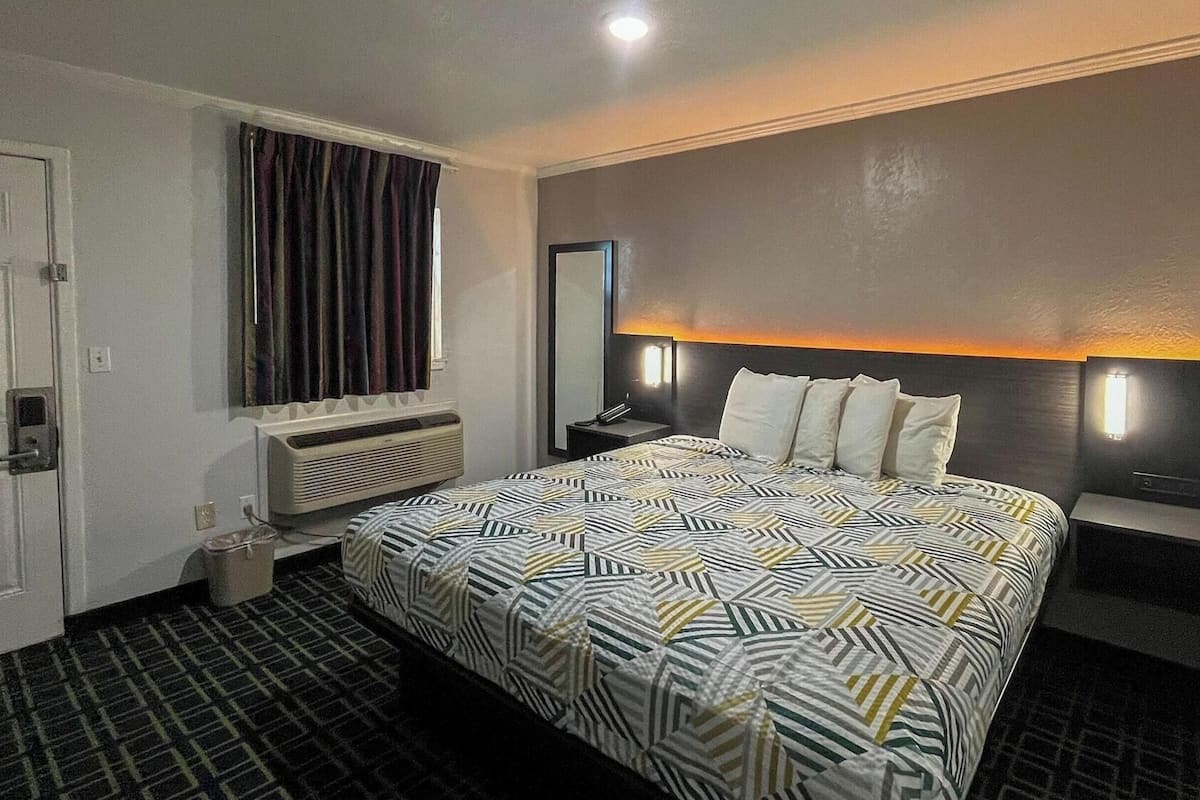 Una habitación de hotel con una cama grande, una mesita de noche, un espejo, una ventana con cortinas y un equipo de aire acondicionado.