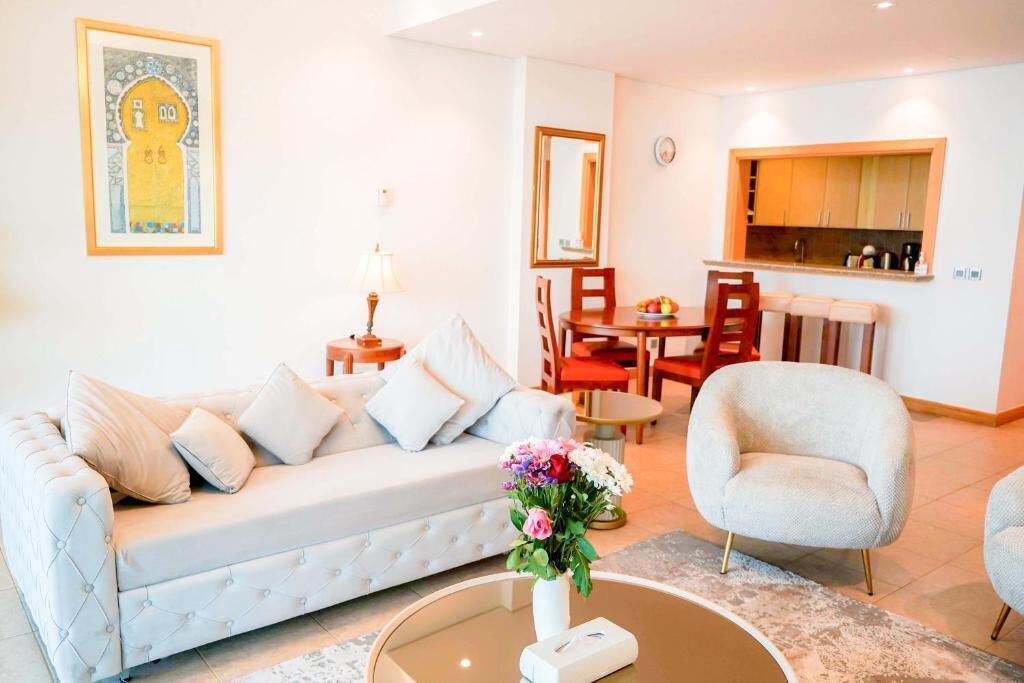 A living room with a sofa, armchair, and a coffee table with a vase of flowers. There is a dining area with a table and chairs, and a kitchen area with cabinets and a window.