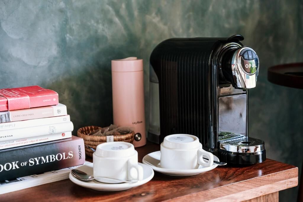 A coffee machine, two cups, a book titled 'Book of Symbols,' and a pink container on a wooden table.