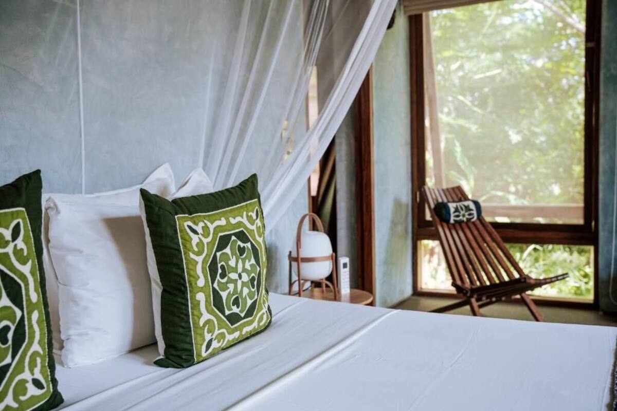 A bed with white and green pillows, a wooden chair, and a window with a view of trees.