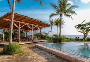 Un espacio al aire libre techado con piscina, palmeras y vista al océano.