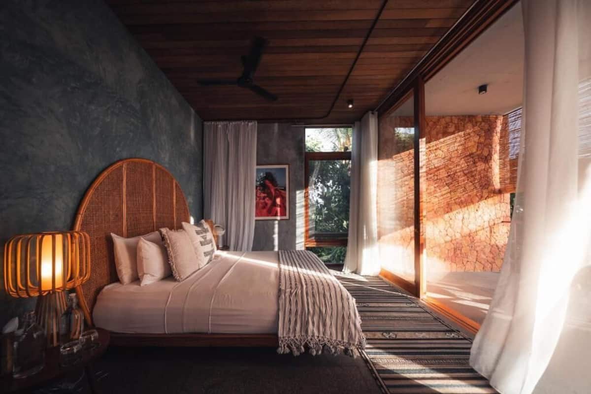 A bedroom with a bed, a nightstand, a lamp, a window, and a view of the outdoors.