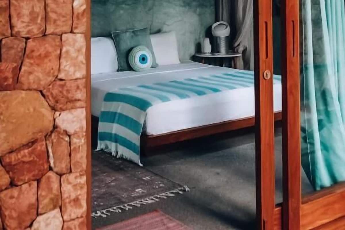A room with a stone wall, a bed with a striped blanket, a dreamcatcher, and a window with curtains.