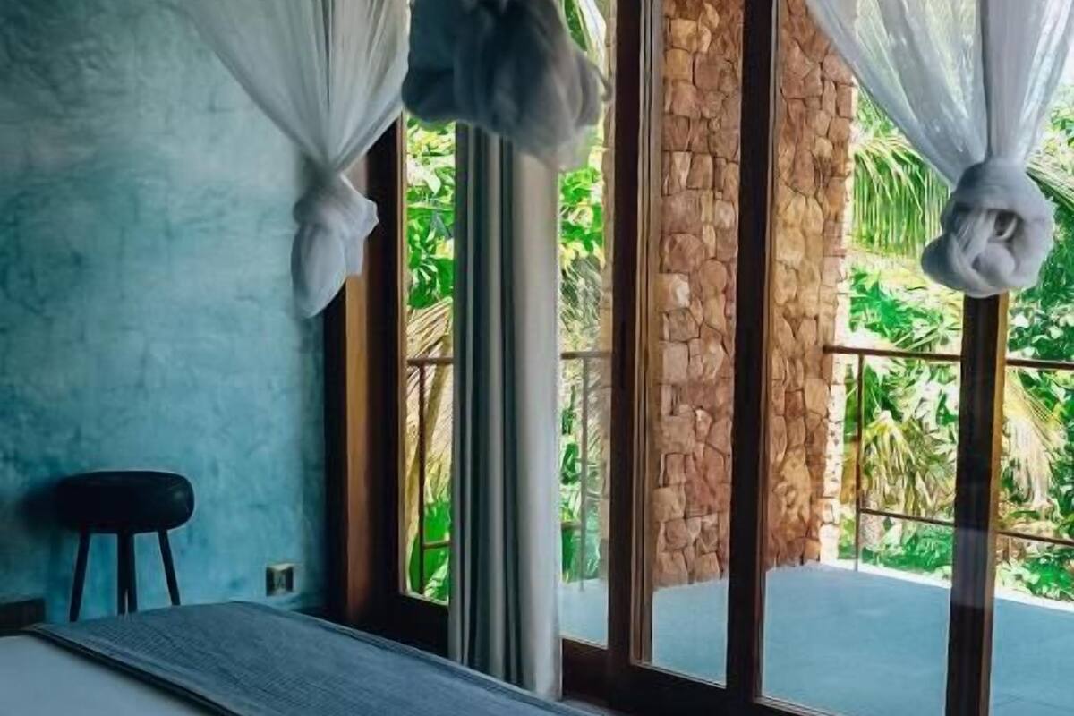 A bedroom with a bed, a stool, and a view of greenery through large windows.
