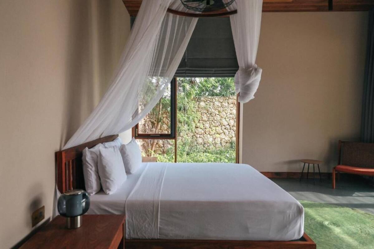 A bedroom with a bed, a bedside table, a chair, and a view of greenery through a window.
