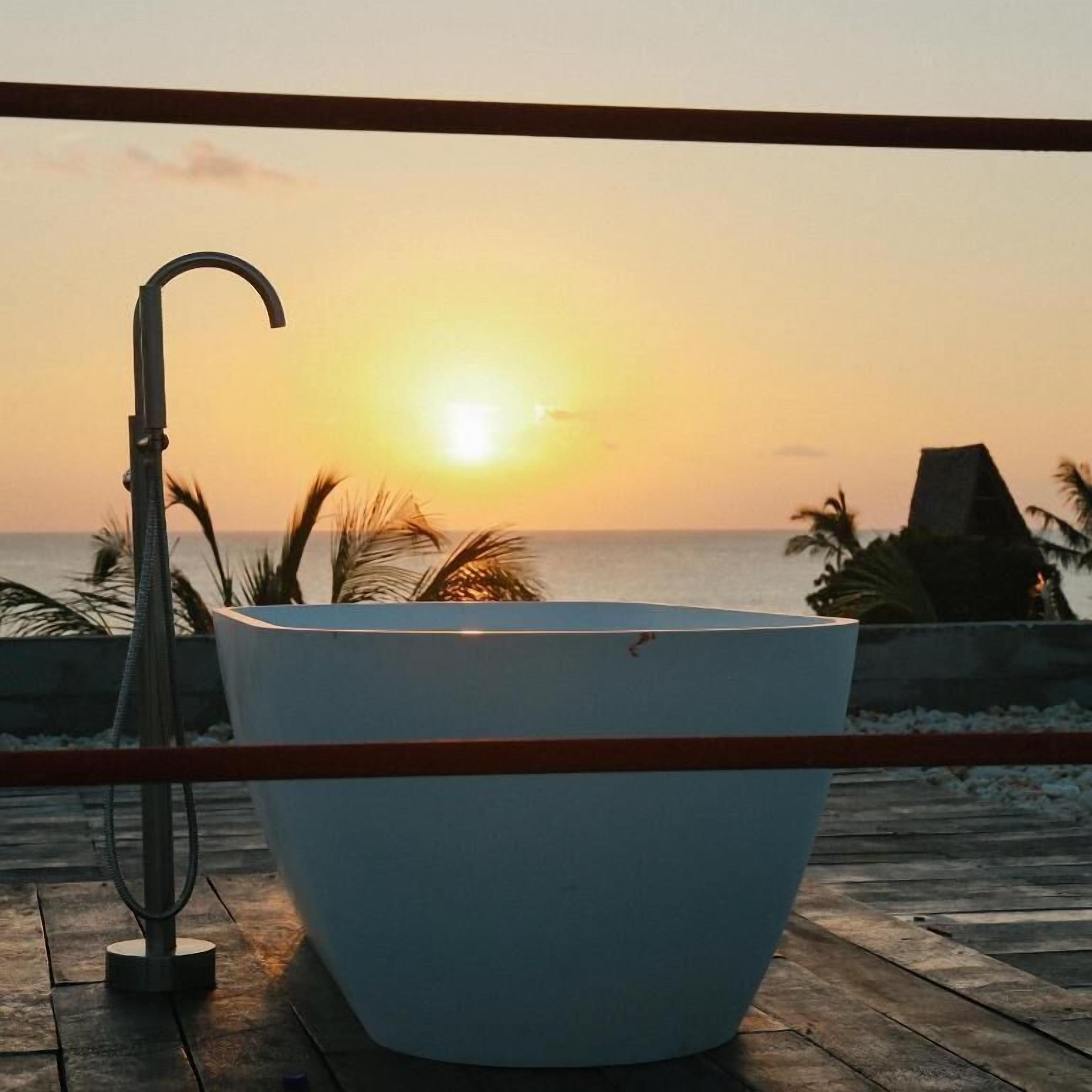 Eine weiße Badewanne auf einer Holzdiele mit Blick auf den Sonnenuntergang über dem Meer.