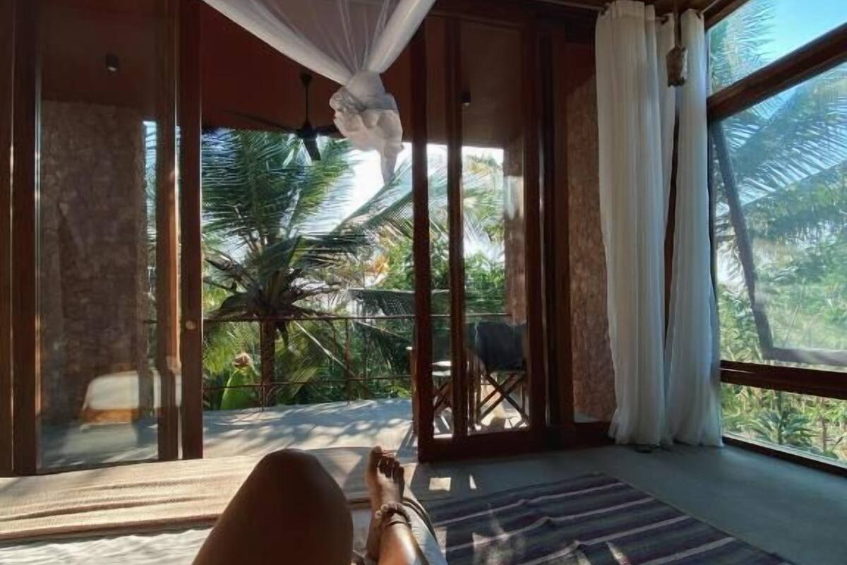 A room with a bed, a view of palm trees, and a large window.