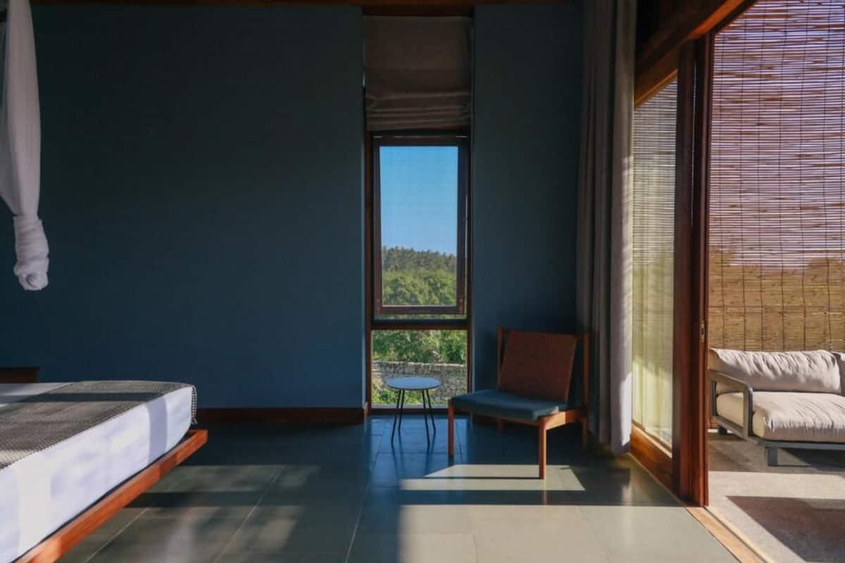 A bedroom with a bed, a chair, a small table, and a view of greenery through a window.