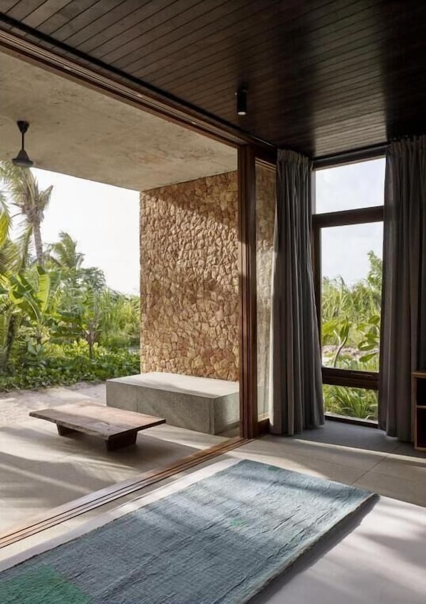 A room with a stone wall, a wooden ceiling, a large window with curtains, a wooden bench, and a view of greenery outside.
