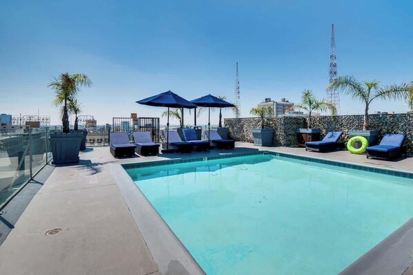 A rooftop pool with sun loungers and umbrellas, surrounded by palm trees and a cityscape view.