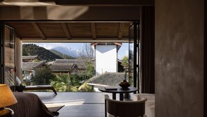 Habitación con vistas a edificios tradicionales y montañas. Se ve una cama, una silla y una mesa con un bol de fruta.