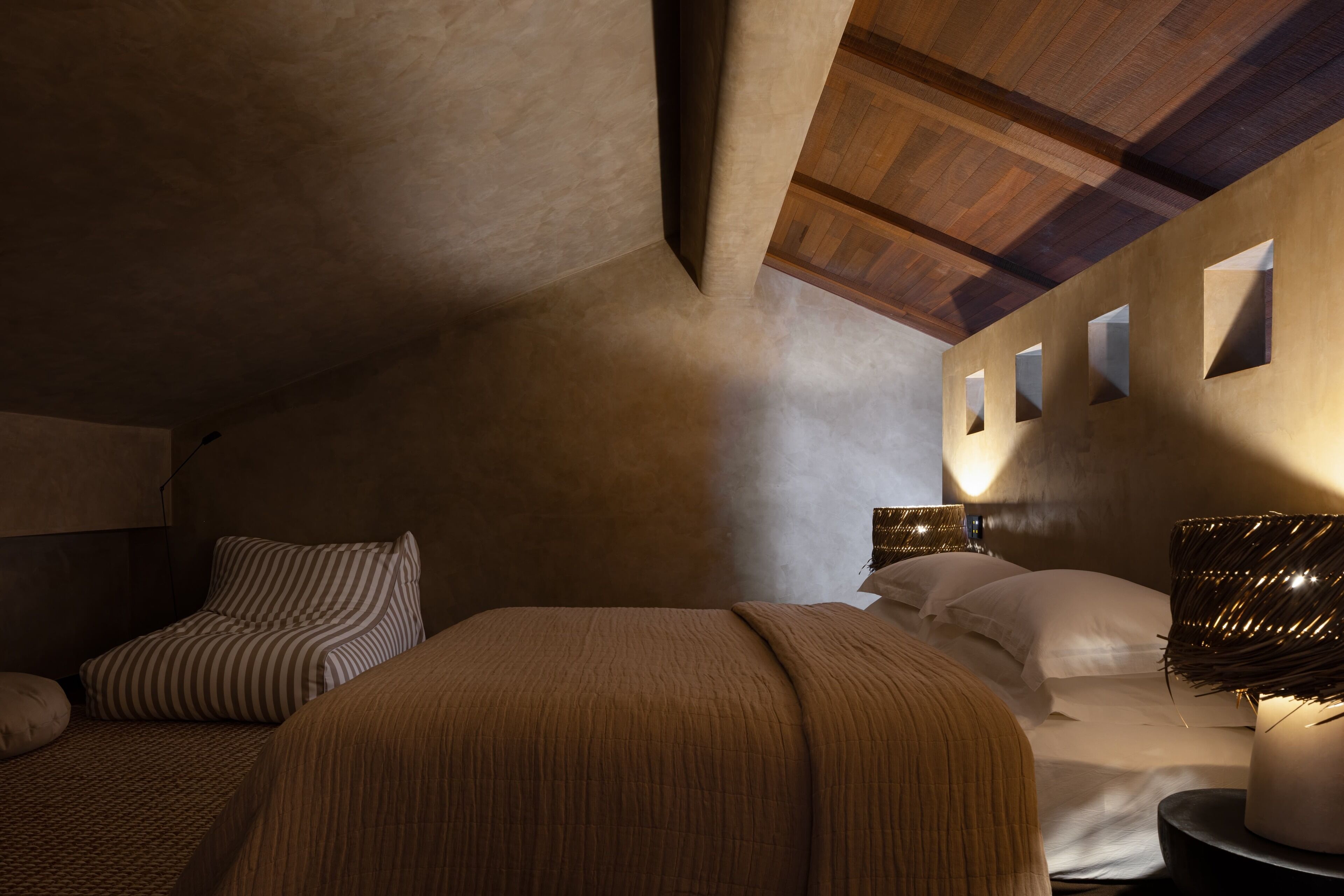 Un dormitorio con techo inclinado, una cama con almohada a rayas, una lámpara de noche y una lámpara de pared.