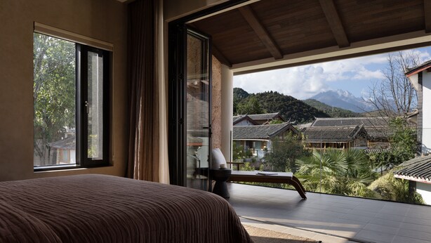Un dormitorio con un ventanal grande, una cama y vistas a edificios tradicionales y montañas.