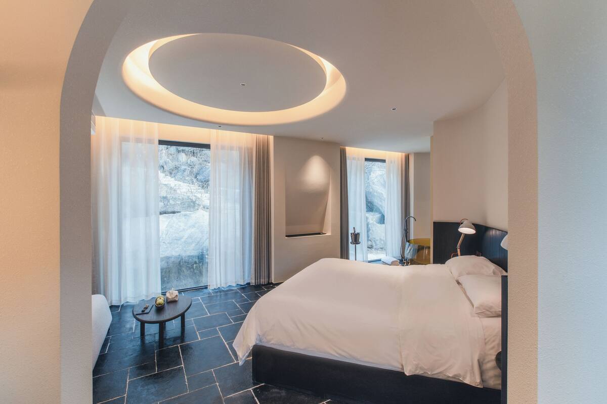 A modern hotel room with a large bed, a small table, and a view of the outdoors through large windows.