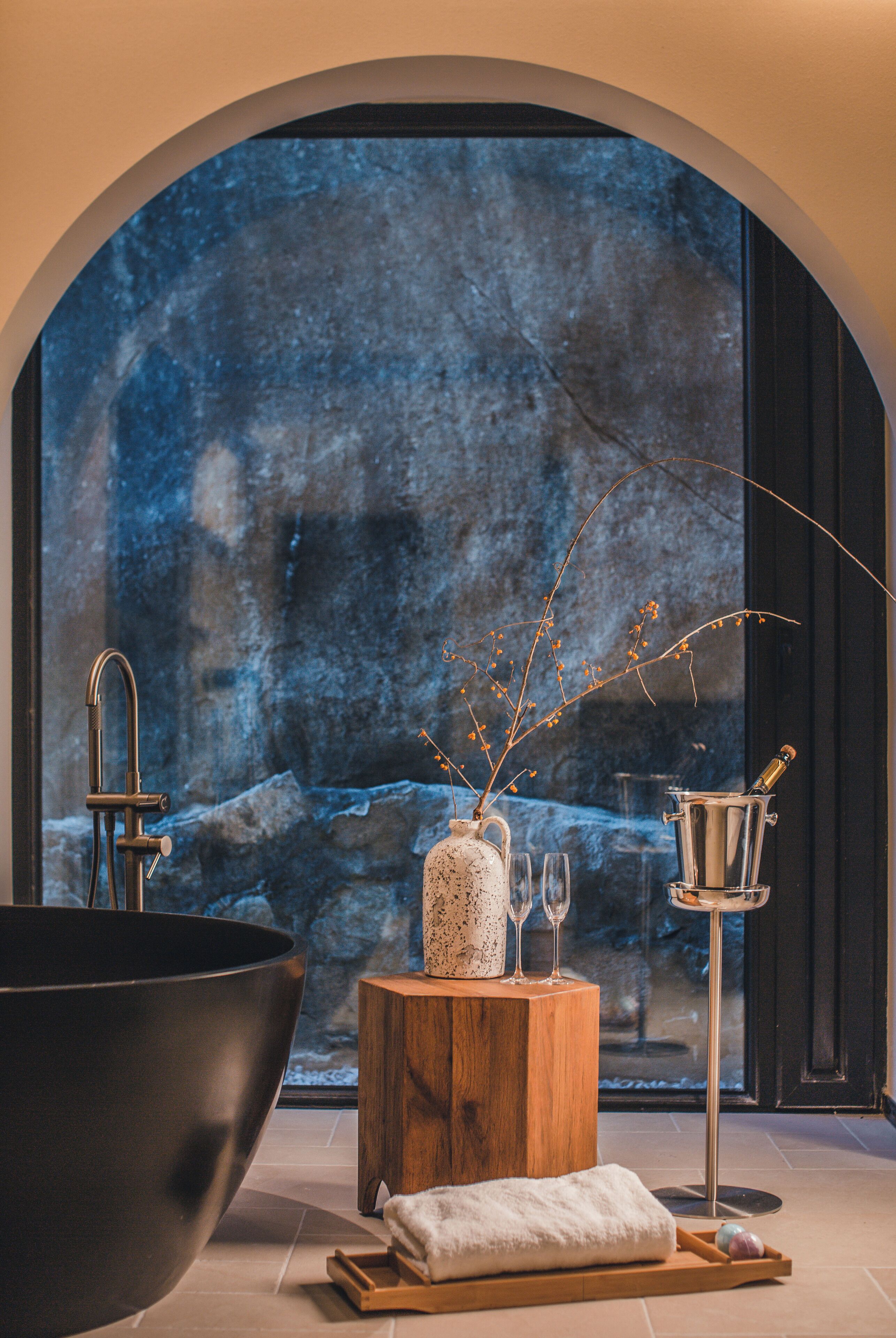 A spa area with a large black bathtub, a wooden stool with a vase and glasses, and a metal container with a bottle.