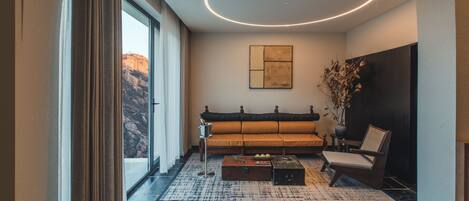 A modern living room with a sofa, a coffee table, and a chair. There is a large window with curtains, a ceiling light, and a wall-mounted artwork.