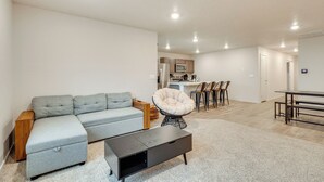 A modern living room with a grey sectional sofa, a black coffee table, and a round chair. A kitchen area with a dining table and chairs is visible in the background.