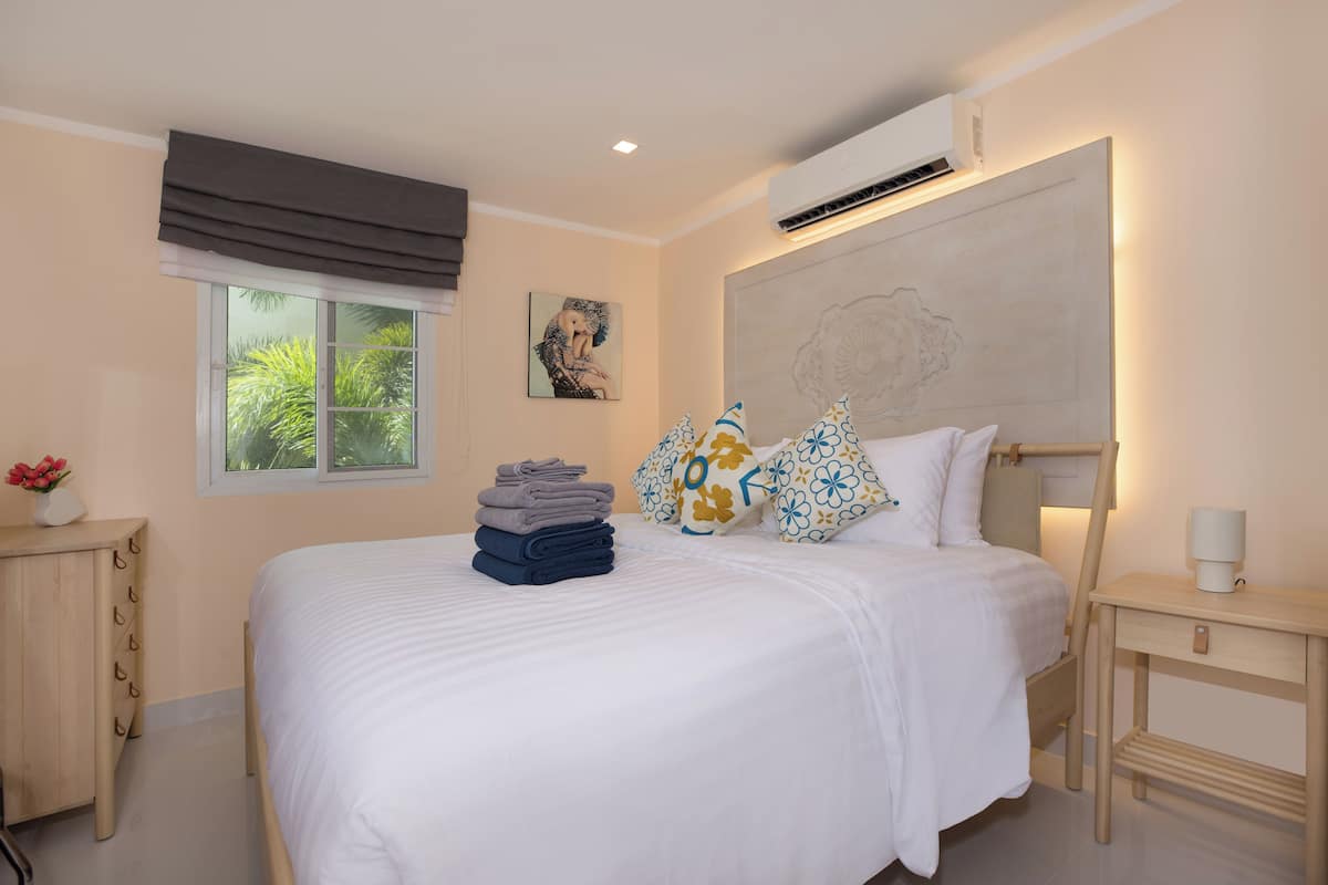 A neatly made bed with folded towels, a bedside table with a lamp, a window with a view of greenery, and a wall-mounted air conditioner.