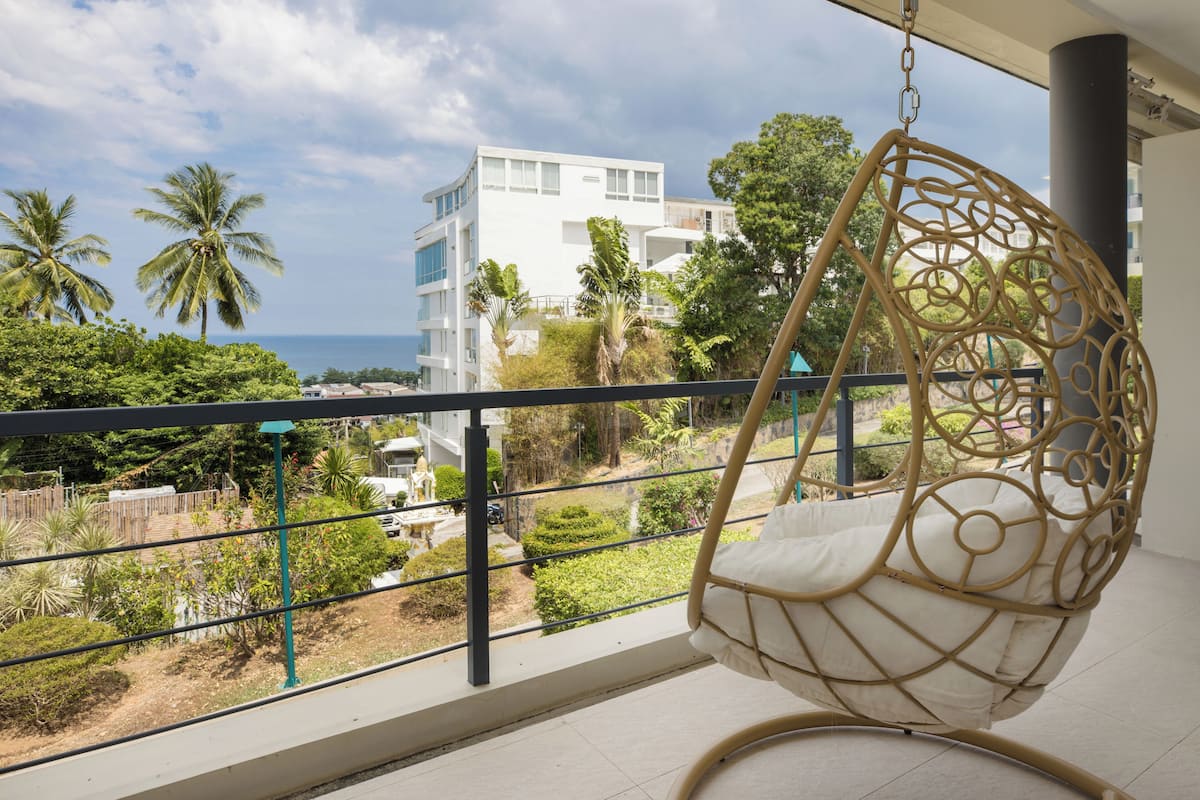A balcony with a hanging chair, a view of a white building, and palm trees.
