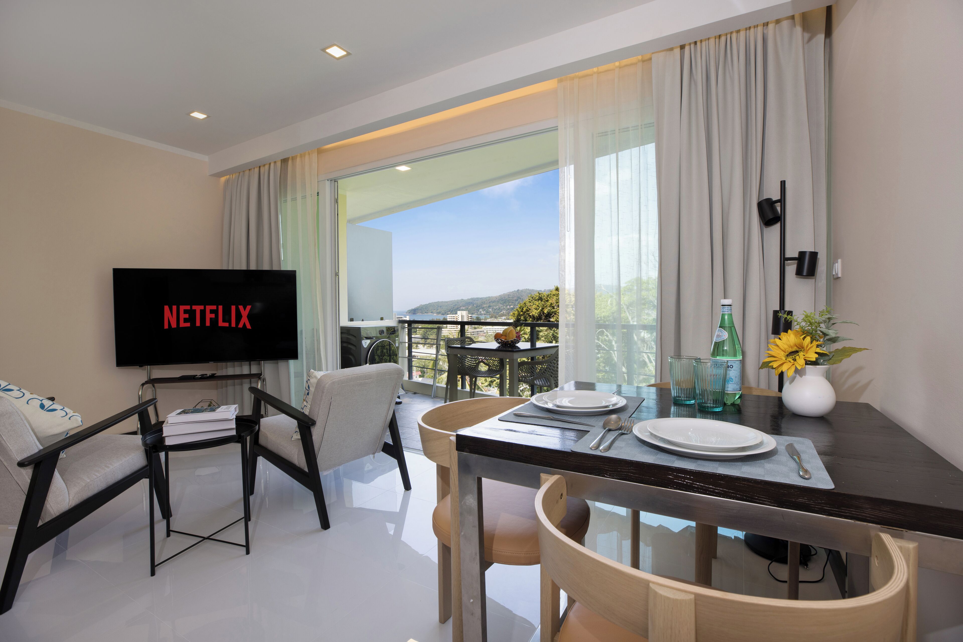 A modern hotel room with a balcony, a dining area with a table set for two, and a flat-screen TV displaying 'NETFLIX'.