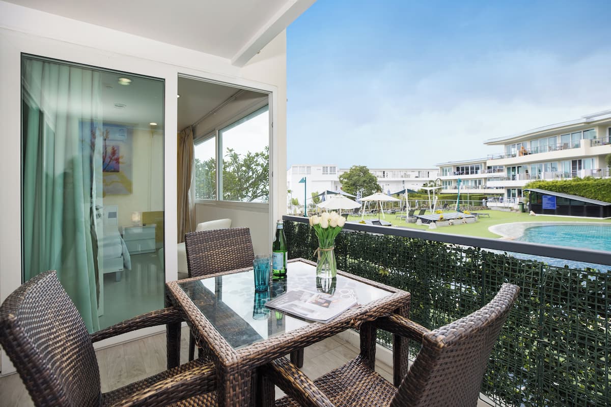 A balcony with wicker furniture, a glass table, and a view of a swimming pool and hotel buildings.