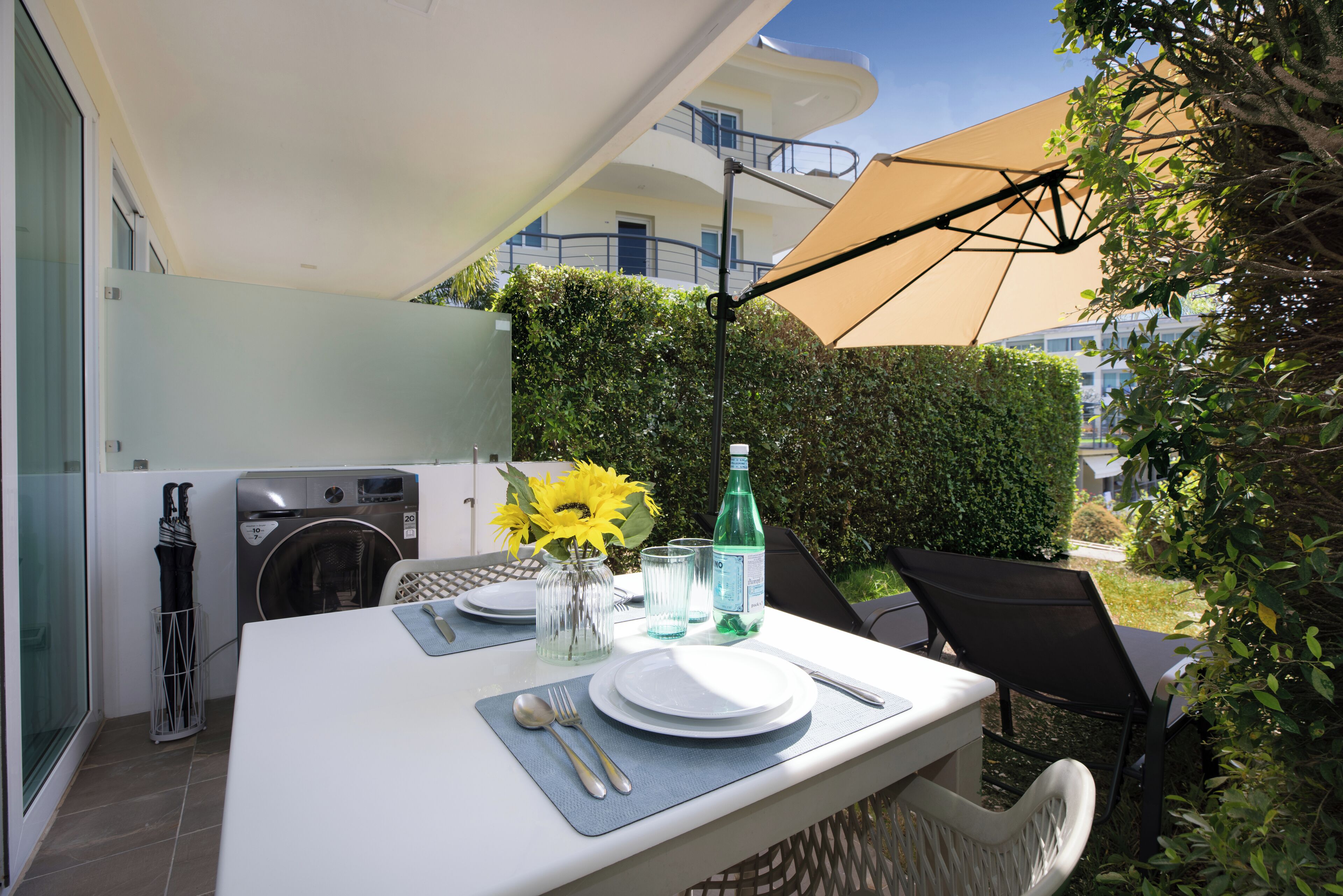 A dining area with a table set for two, a washing machine, and a large umbrella providing shade.