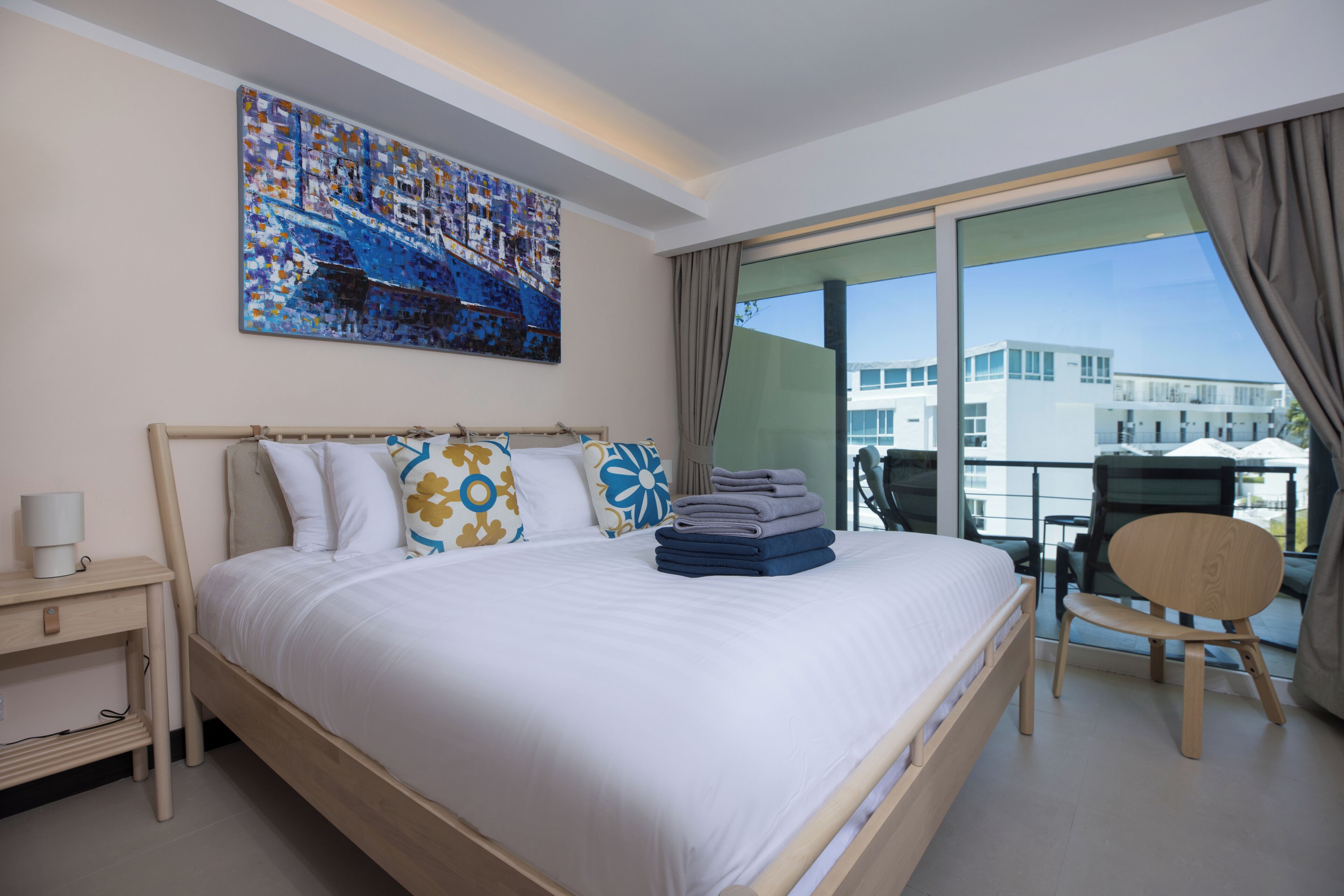 Habitación de hotel con cama, escritorio, silla, balcón con vista a edificios y un cuadro en la pared.