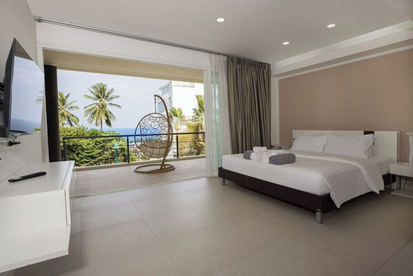 A hotel room with a bed, a TV, a balcony with a view of palm trees and the ocean, and a chair. - Woodbridge Property at Karon Butterfly (Karon)