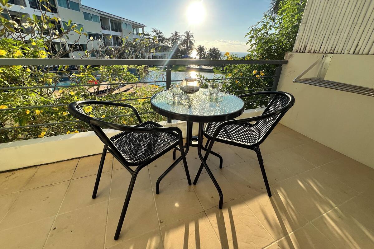 A balcony with a round glass table and two chairs, overlooking a pool and buildings.