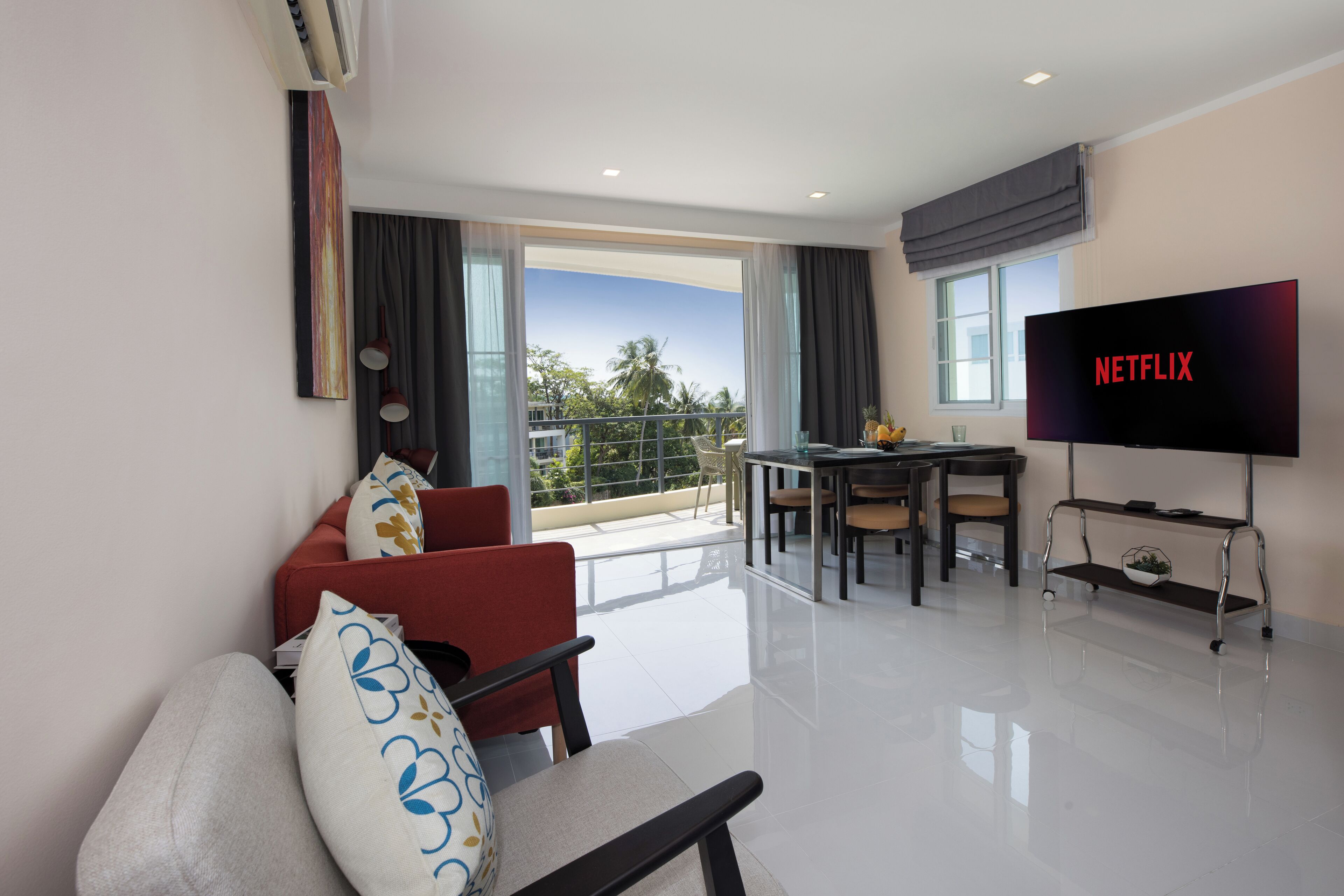 Una sala moderna con una televisión de pantalla plana mostrando "NETFLIX", un comedor con mesa y sillas, y un balcón con vistas a palmeras.