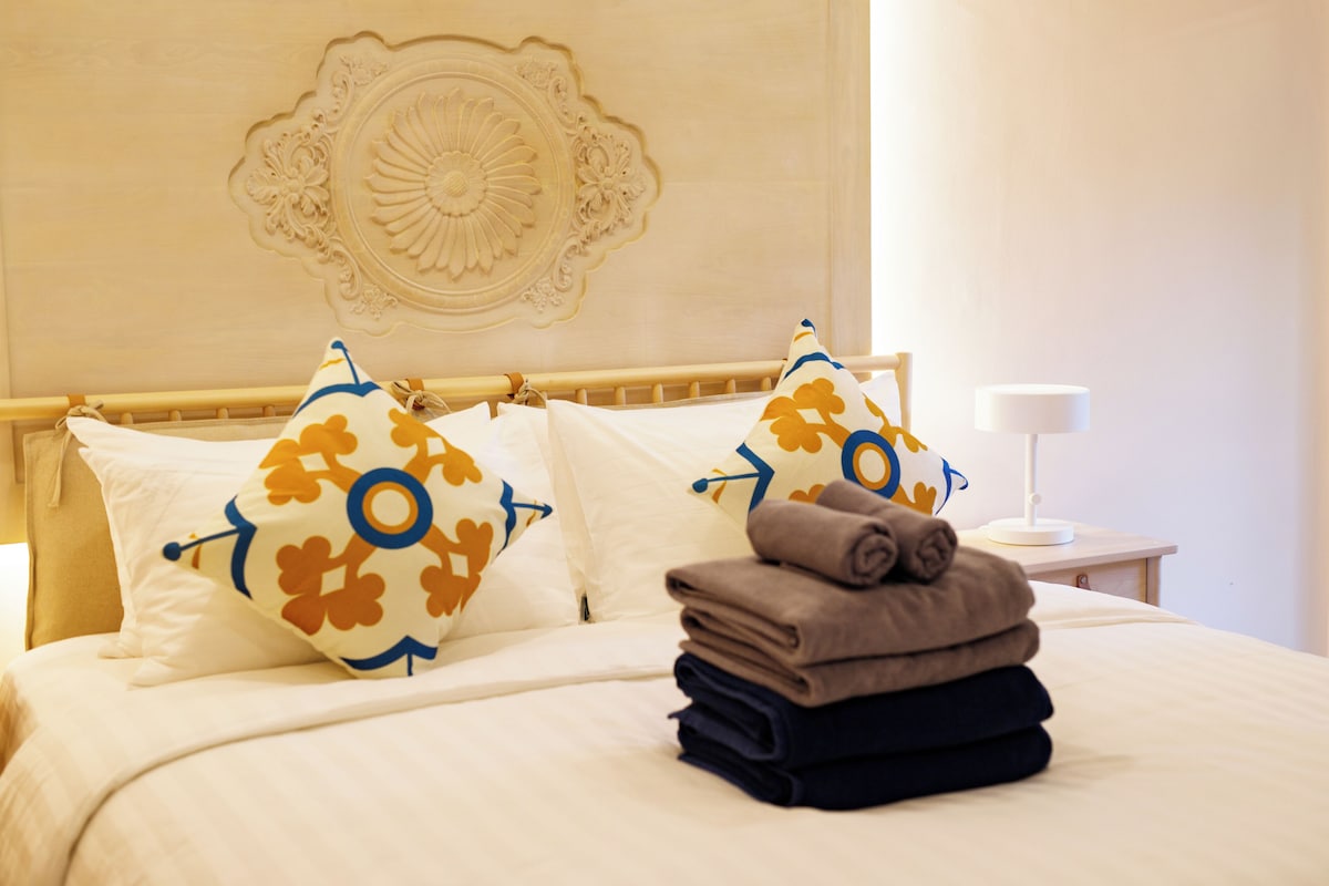 A neatly made bed with patterned pillows, a folded towel stack, and a decorative wall piece.