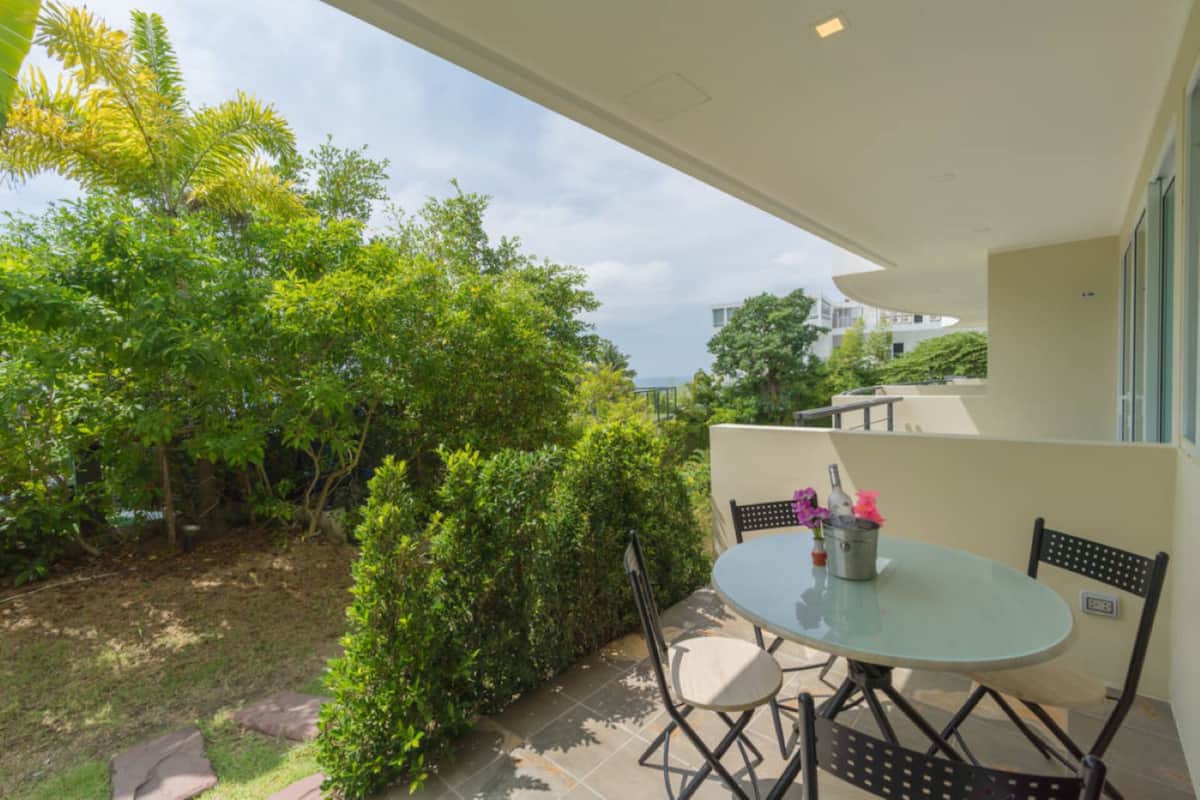 An outdoor patio with a round glass table and chairs, surrounded by greenery and a view of a building in the background.