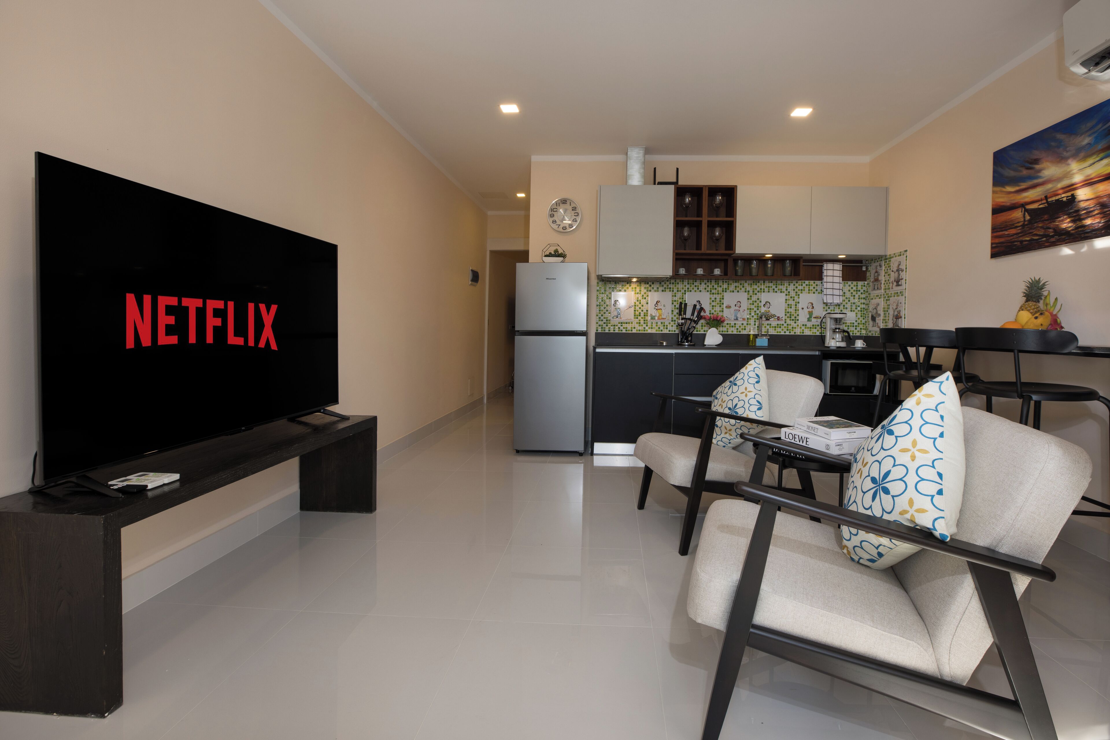 En moderne stue med et fladskærms-TV, der viser 'NETFLIX', et køkken med et køleskab, og et spiseområde med stole og et bord.