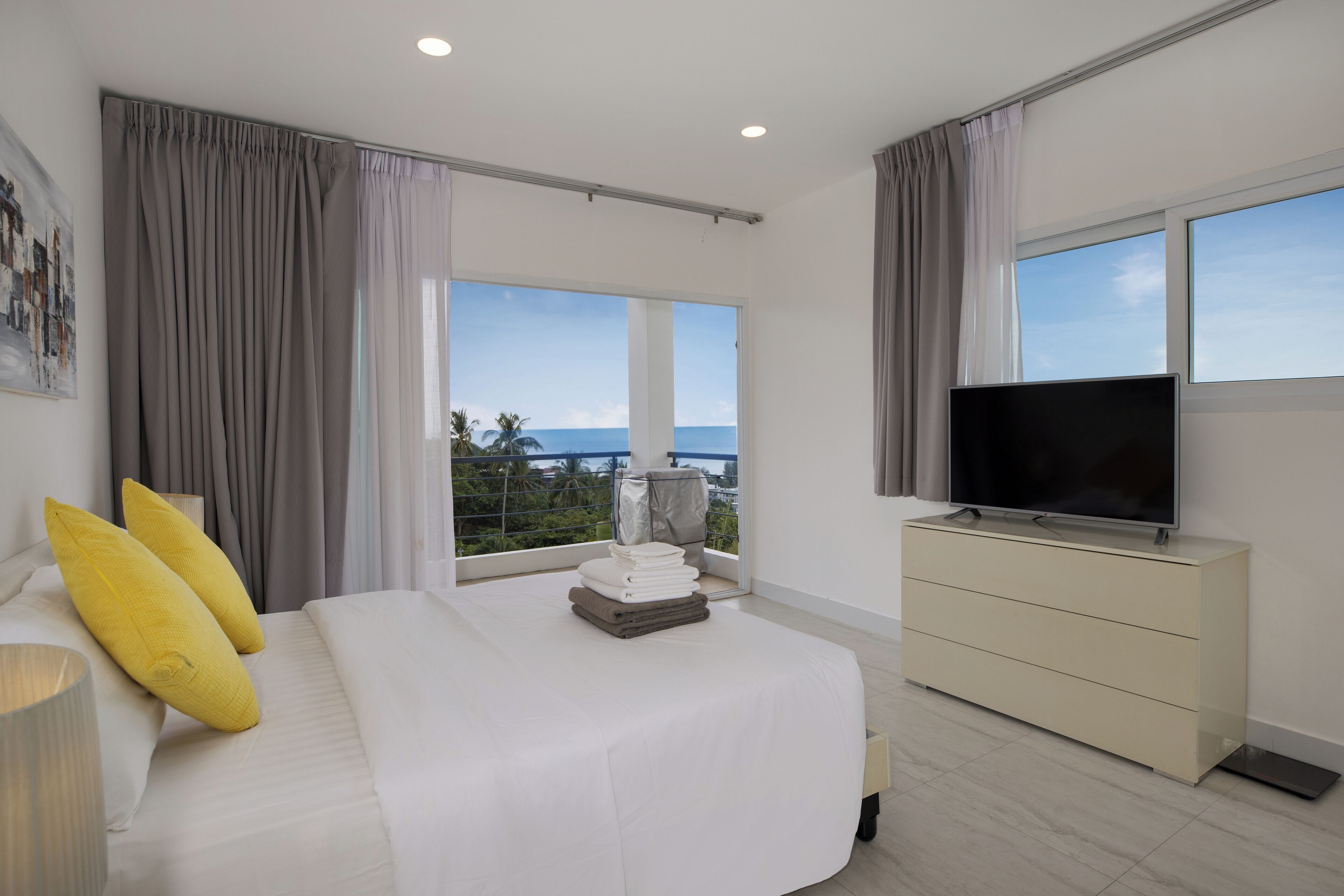 A modern hotel room with a large bed, a flat-screen TV, a dresser, and a balcony with a view of the ocean.