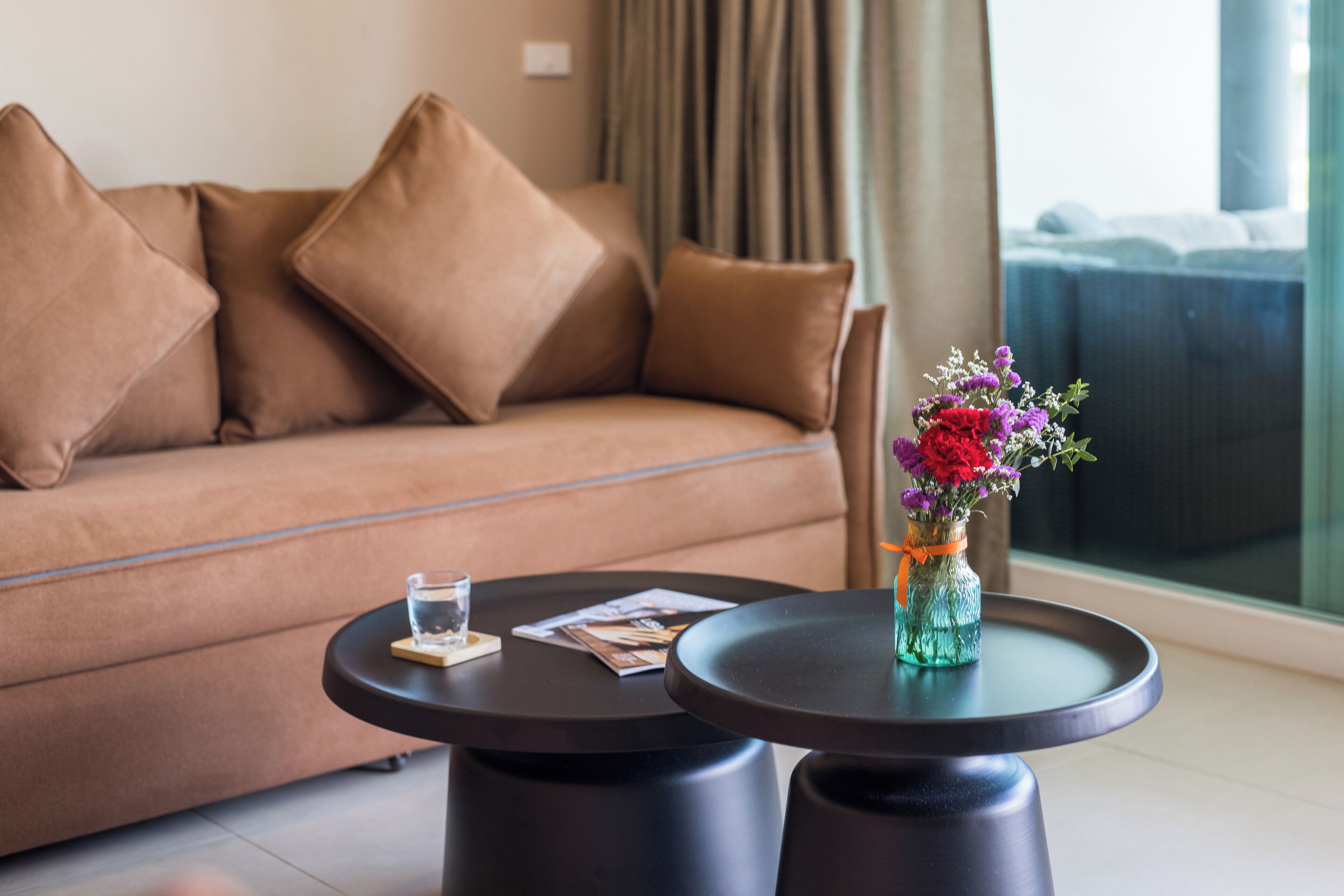 Un salon moderne avec un canapé marron, deux petites tables rondes noires, un verre d’eau, un magazine et un vase rempli de fleurs.