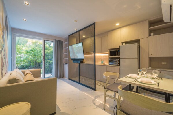 A modern kitchen with a dining area, a TV mounted on a partition, and a view of greenery outside. - Woodbridge Property at Karon Butterfly (Karon)
