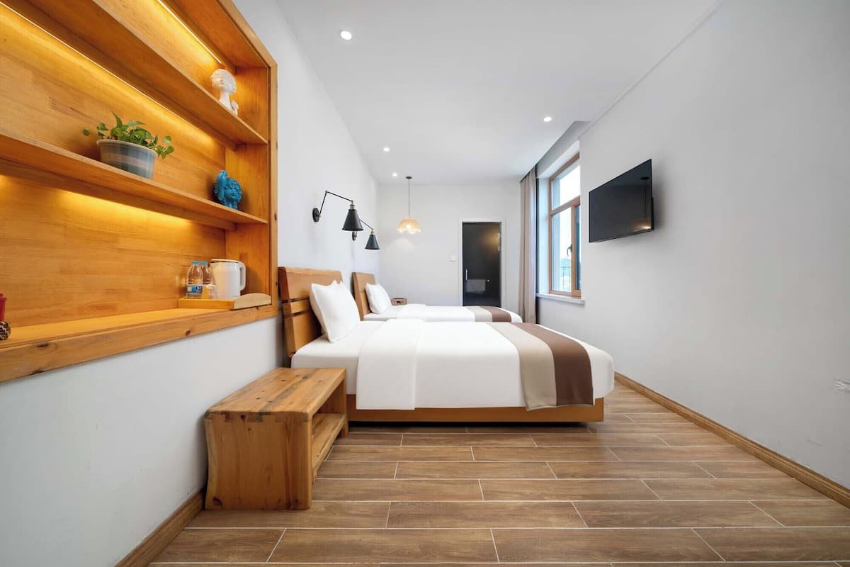 A modern hotel room with a wooden bench, a bed with white bedding, a wooden shelf with decorative items, a wall-mounted TV, and a window providing natural light.
