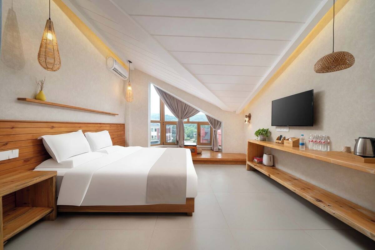 A modern hotel room with a wooden bed, a TV mounted on the wall, and a large window offering a view of the outside.