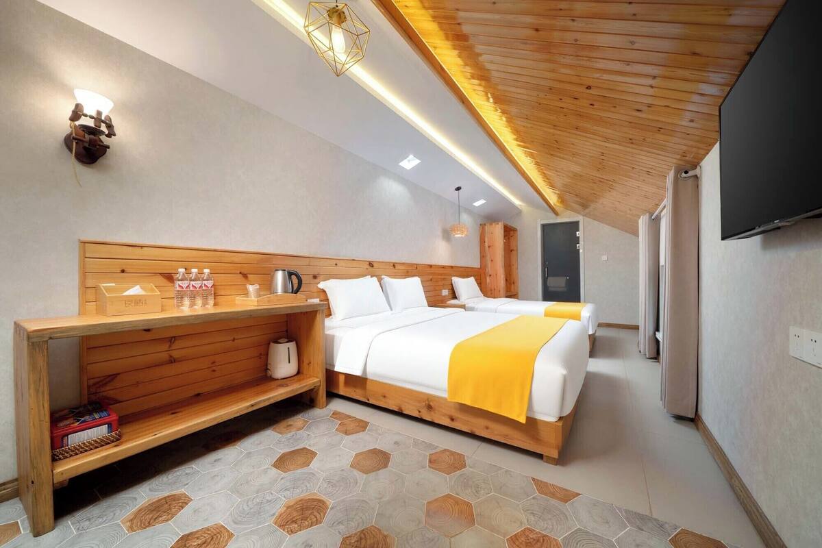 A modern hotel room with a wooden headboard, a bed with a yellow blanket, a bedside table with a kettle and bottles, and a TV on the wall.