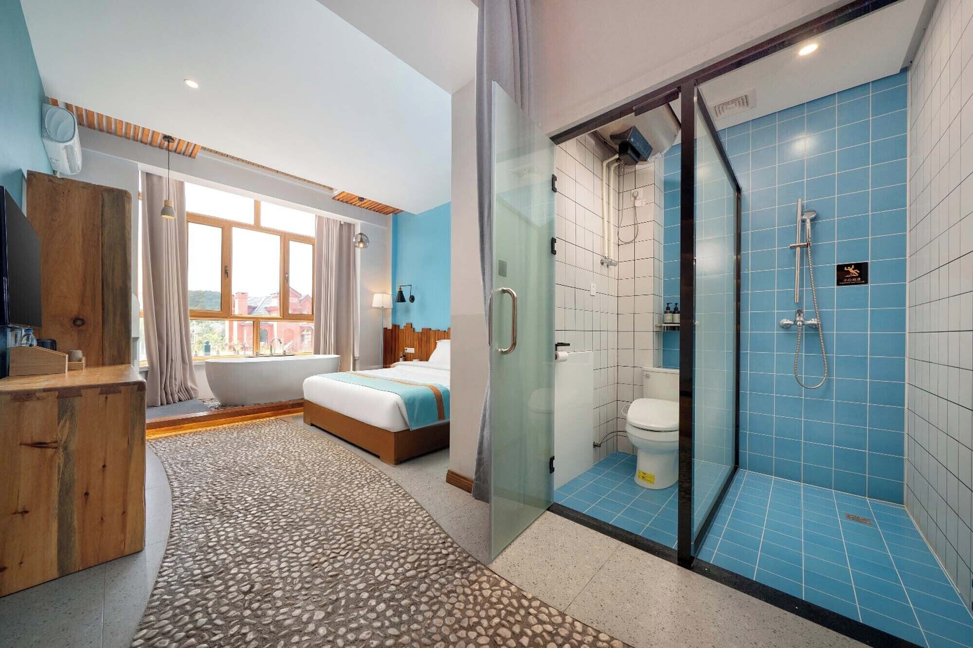 A modern hotel room with a large bed, a shower with blue tiles, and a view of buildings outside the window.
