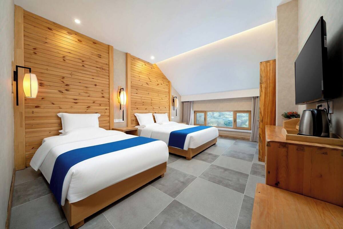 A hotel room with two beds, wooden panel walls, a large window, and a flat-screen TV.